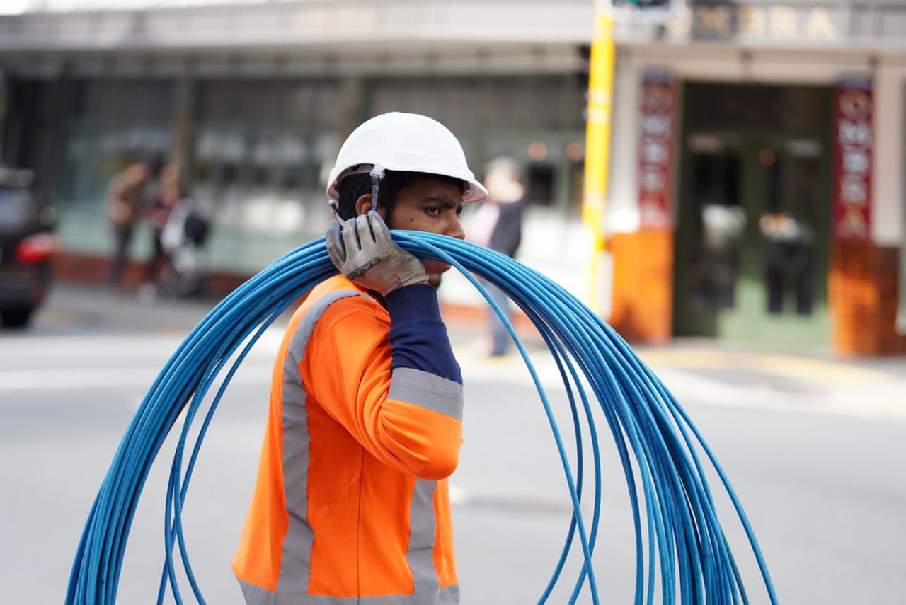 Construction worker in Wellington carrying blue cables, wearing safety gear on a city street.